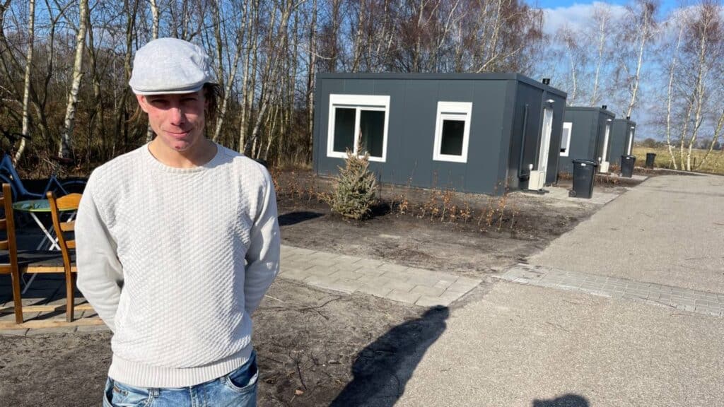 Jonge man met een wit petje en een witte trui, met op de achtergrond een paar kleine woningen.
